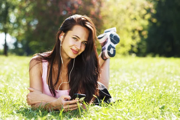 Young woman in dress lies on green grass and listens to music in — Stock Photo, Image