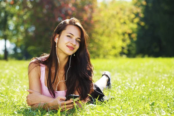 Young woman in dress lies on green grass and listens to music in — Stock Photo, Image