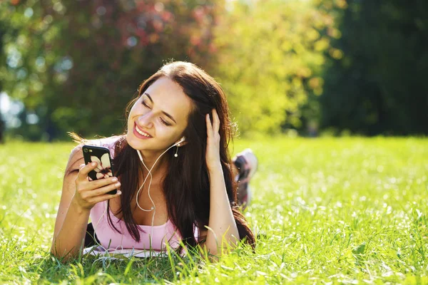 Young woman in dress lies on green grass and listens to music in — Stock Photo, Image