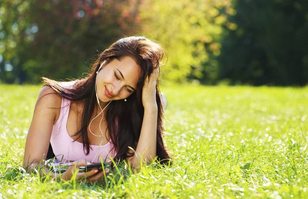 Young woman in dress lies on green grass and listens to music in — Stock Photo, Image