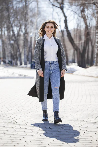 Chica morena en un abrigo gris y pantalones vaqueros azules camina en el parque de primavera —  Fotos de Stock