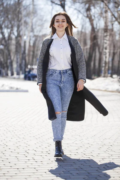 Brünettes Mädchen in grauem Mantel und blauer Jeans spaziert im Frühlingspark — Stockfoto