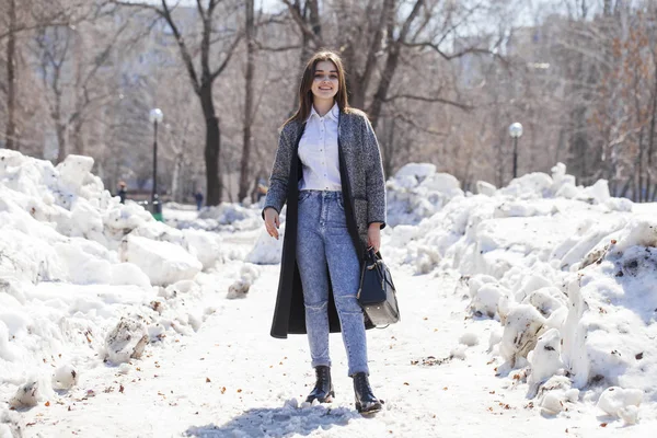 Брюнетка в сером пальто и синих джинсах гуляет по весеннему парку — стоковое фото