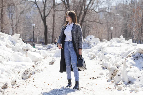 Chica morena en un abrigo gris y pantalones vaqueros azules camina en el parque de primavera — Foto de Stock