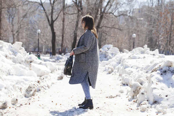 Menina morena em um casaco cinza e jeans azuis caminha no parque da primavera — Fotografia de Stock