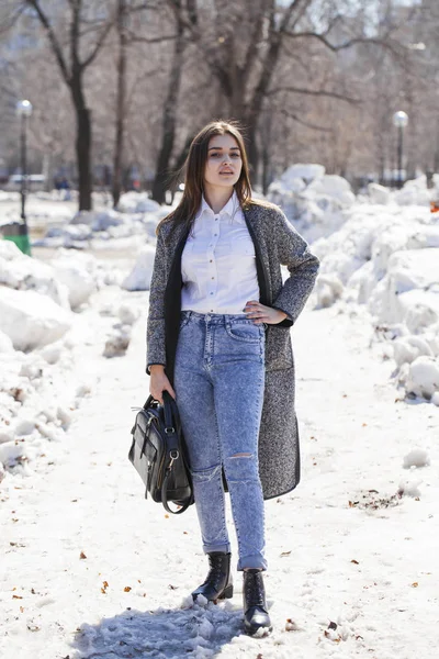 Брюнетка в сером пальто и синих джинсах гуляет по весеннему парку — стоковое фото