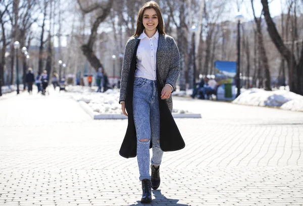 Gadis berambut cokelat dengan mantel abu-abu dan celana jeans biru berjalan di taman musim semi — Stok Foto