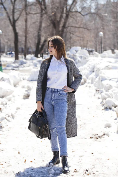 Brunette girl in a gray coat and blue jeans walks in spring park — Stock Photo, Image