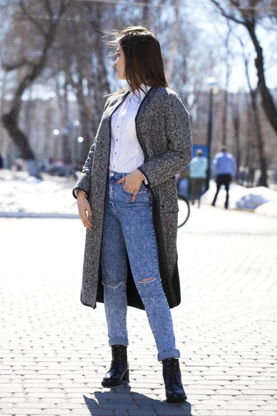 Chica morena en un abrigo gris y pantalones vaqueros azules camina en el parque de primavera —  Fotos de Stock