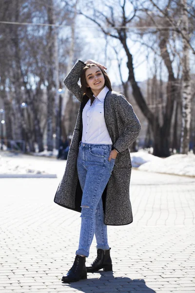 Brunette girl in a gray coat and blue jeans walks in spring park — Stock Photo, Image