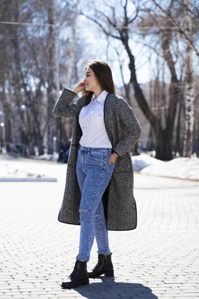 Brünettes Mädchen in grauem Mantel und blauer Jeans spaziert im Frühlingspark — Stockfoto