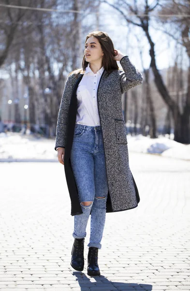 Brünettes Mädchen in grauem Mantel und blauer Jeans spaziert im Frühlingspark — Stockfoto