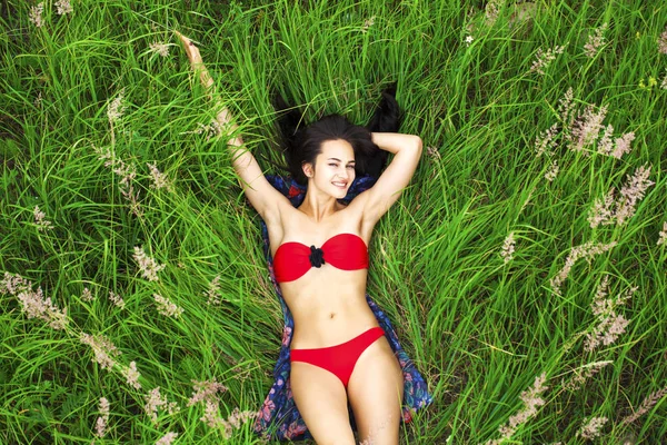 Sexy brunette woman in a red bathing suit lying on the grass top — Stock Photo, Image