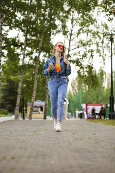 年轻的金发女郎穿蓝色牛仔裤走在夏天公园 — 图库照片