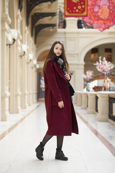 Vacker flicka i stilfull i en lång vinröd kappa, inomhus shop — Stockfoto