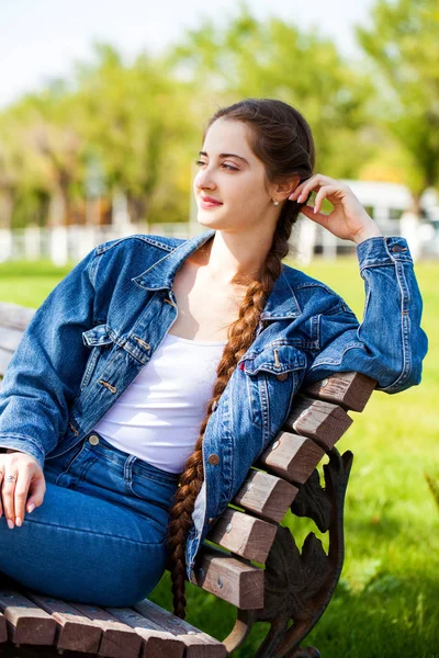 Krásná dívka v džínové bundě sedí na lavičce v létě par — Stock fotografie