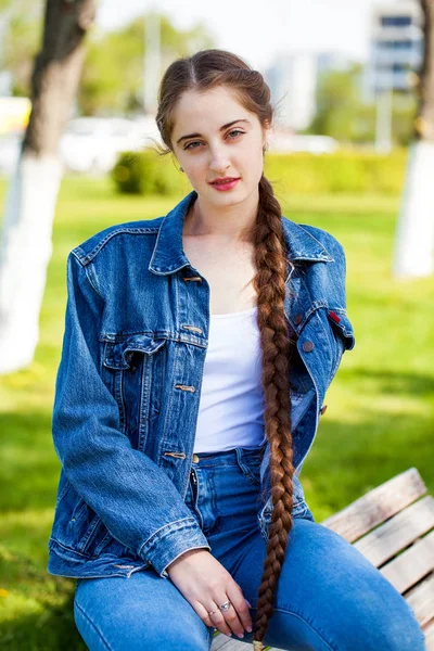 Hermosa chica en una chaqueta de mezclilla se sienta en un banco en un par de verano —  Fotos de Stock