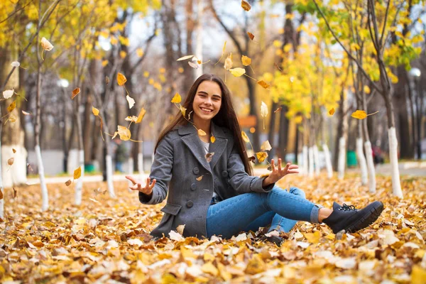 Ung flicka i blå jeans och grå rock sitter på höstlöv i — Stockfoto