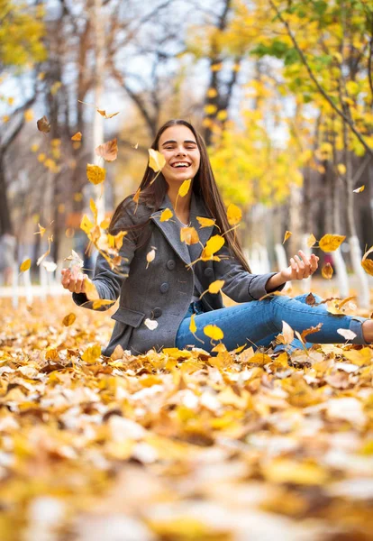 Chica joven en pantalones vaqueros azules y abrigo gris se sienta en las hojas de otoño en — Foto de Stock