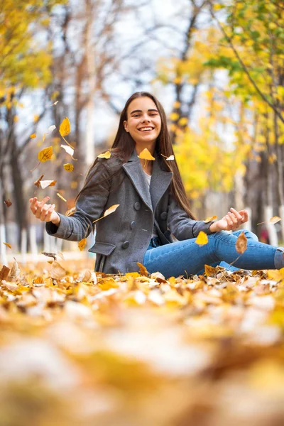 Молодая девушка в синих джинсах и сером пальто сидит на осенних листьях в — стоковое фото