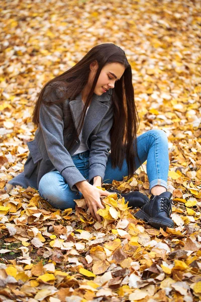 Menina jovem em jeans azul e casaco cinza senta-se nas folhas de outono em — Fotografia de Stock