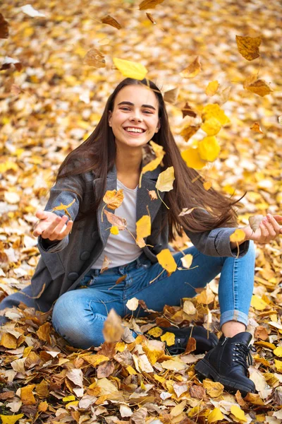 Young girl in blue jeans and gray coat sits on autumn leaves in — Stock Photo, Image