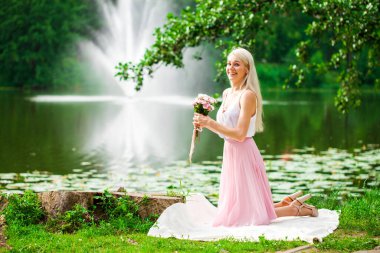 Bir yaz parkında gölün kenarında dinlenen genç bir kadının tam boy portresi.