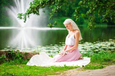 Bir yaz parkında gölün kenarında dinlenen genç bir kadının tam boy portresi.