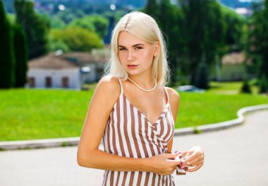 Çizgili bir yaz elbisesi içinde, güneşli bir havada sokakta poz veren genç, güzel bir sarışın.