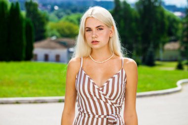 Çizgili bir yaz elbisesi içinde, güneşli bir havada sokakta poz veren genç, güzel bir sarışın.