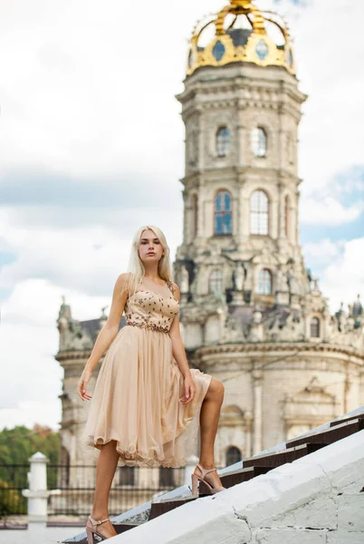 Full body portrait of a young beautiful blonde woman in beige dress against the background of the Temple of the Sign of the Most Holy Mother of God in Dubrovitsy