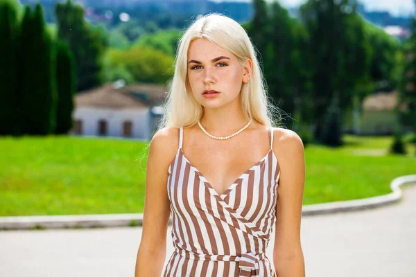 Çizgili bir yaz elbisesi içinde, güneşli bir havada sokakta poz veren genç, güzel bir sarışın.