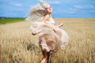 Moda elbisesi giymiş, buğday tarlasında yürüyen güzel bir sarışının vücut portresi.