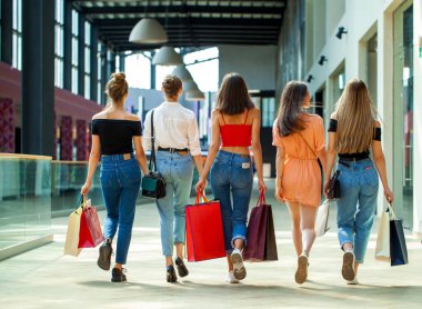 Kadın alışverişi. Arkaya bakan genç kızlar alışveriş merkezinde çantalarla yürüyorlar.