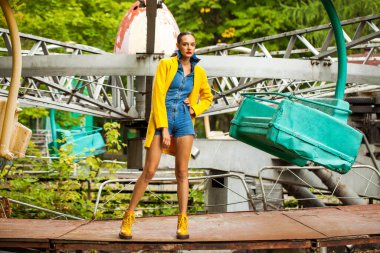 Bir lunaparkta atlıkarınca arka planında poz veren sarı ceketli ve kot pantolonlu manken.