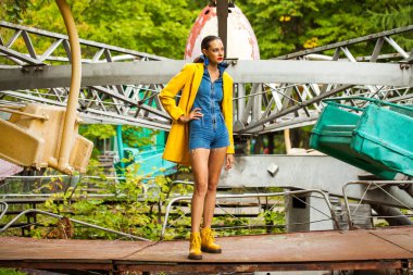 Bir lunaparkta atlıkarınca arka planında poz veren sarı ceketli ve kot pantolonlu manken.