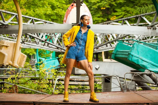 遊園地のカルーセルを背景にした黄色のコートとデニムのオーバーオールのファッションモデル — ストック写真