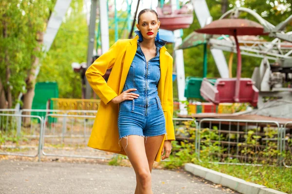 遊園地のカルーセルを背景にした黄色のコートとデニムのオーバーオールのファッションモデル — ストック写真