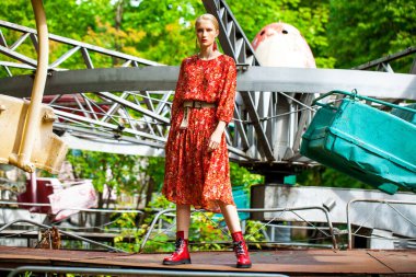 Uzun kırmızı çiçekli şık bir modelin tüm vücut portresi eğlence parkının arka planında poz veriyor. 