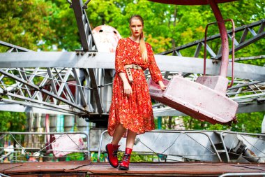 Uzun kırmızı çiçekli şık bir modelin tüm vücut portresi eğlence parkının arka planında poz veriyor. 