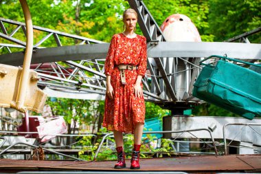 Uzun kırmızı çiçekli şık bir modelin tüm vücut portresi eğlence parkının arka planında poz veriyor. 