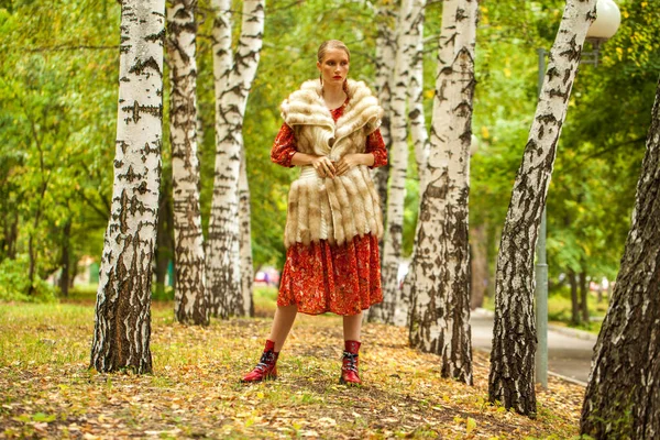 Russian beauty woman. Full body portrait of a stylish model in long red flowers dress and fur vest posing in autumn forest