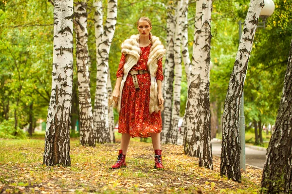 Russian beauty woman. Full body portrait of a stylish model in long red flowers dress and fur vest posing in autumn forest