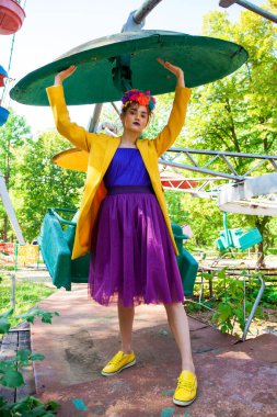 Sarı ceketli şık bir mankenin portresi lunaparkın arka planında poz veriyor. 