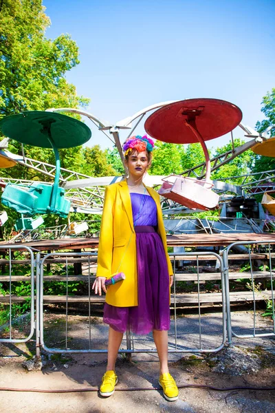 Sarı ceketli şık bir mankenin portresi lunaparkın arka planında poz veriyor. 