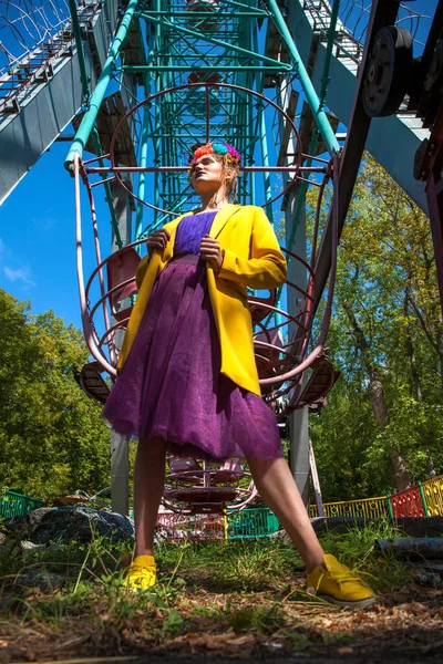 Sarı ceketli şık bir mankenin portresi lunaparkın arka planında poz veriyor. 