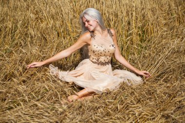 Moda elbisesi giymiş, buğday tarlasında yürüyen güzel bir sarışının vücut portresi.