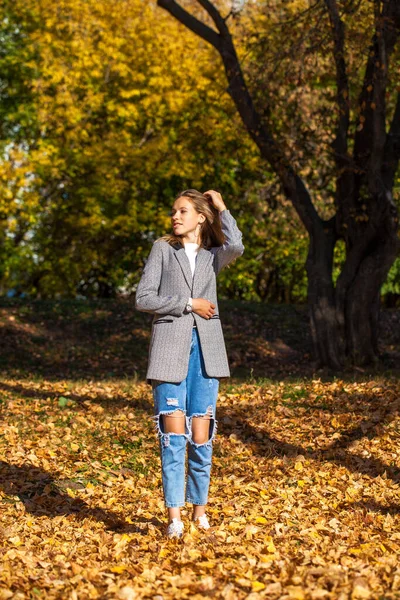 一个穿着时髦撕破牛仔裤的年轻漂亮姑娘在秋天的公园里摆姿势的全长肖像 — 图库照片