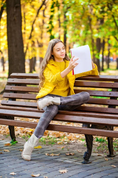 若いです美しいです女の子は秋の公園に座っている間スケッチブックに鉛筆で描きます — ストック写真