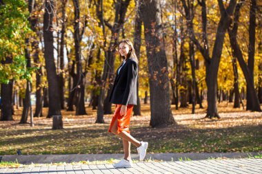 Sonbahar parkında yürüyen turuncu etekli esmer bir kadının vücut portresi.